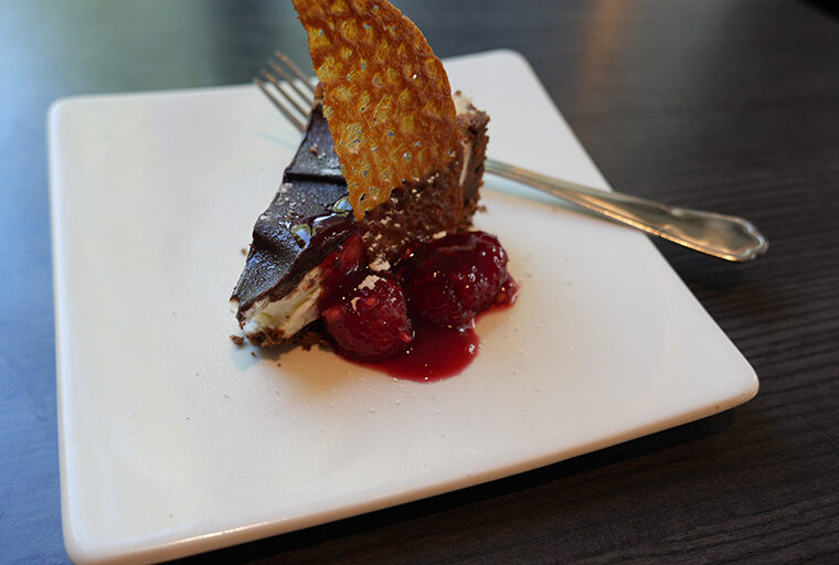 A piece of chocolate dessert sat on a white square plate with a fork resting to one side